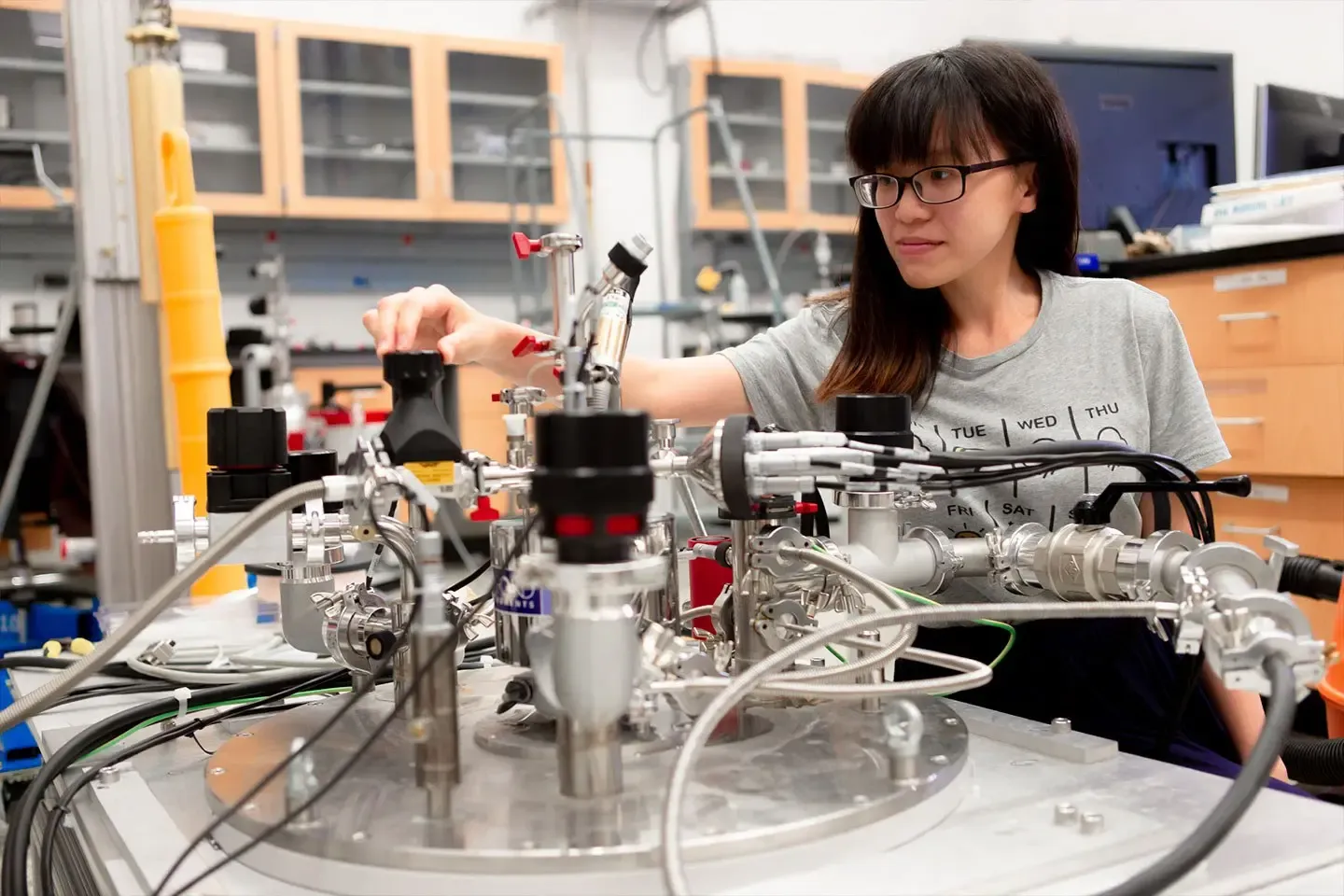 A technician stands behind a large test chamber while adjusting dials. Many cables, and tubing wind in and around the equipment