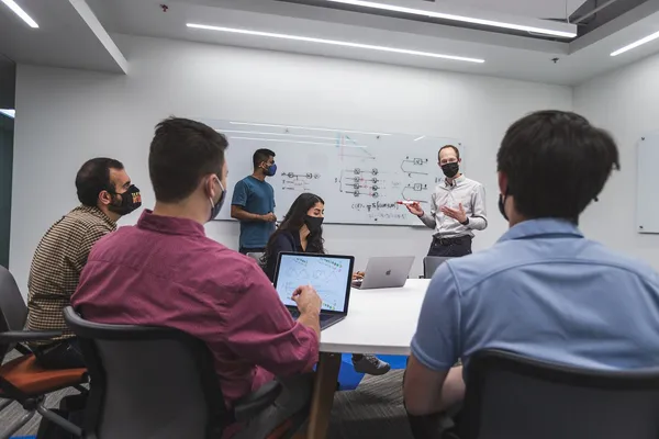 A number of individuals collaborate to solve a problem. Some are in front of a dry erase board, and some individuals are sitting at a table with notebook computers. Everyone is wearing face masks to protect against COVID.
